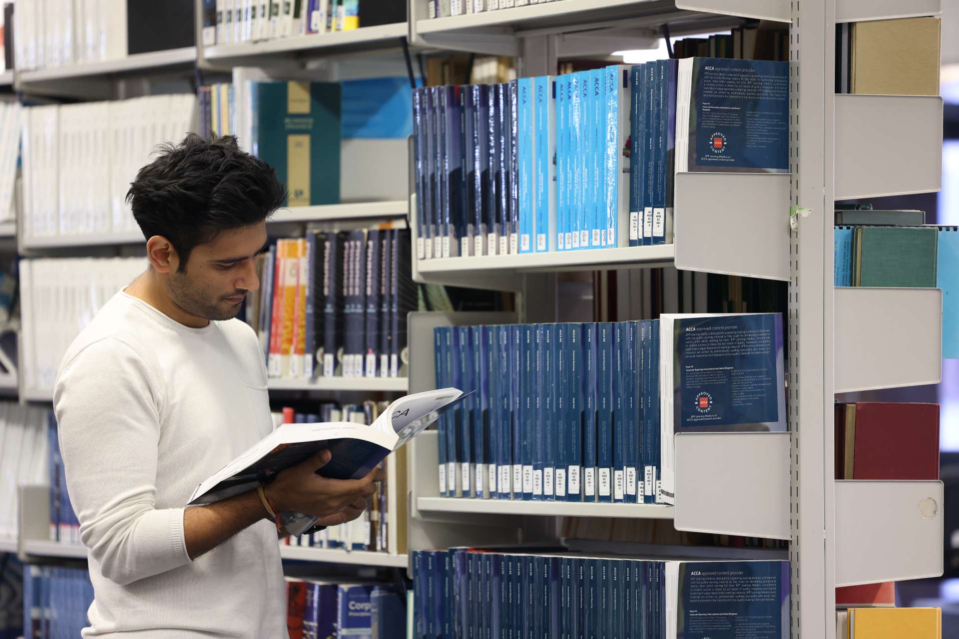 A postgraduate student standing in the library, reading a book.