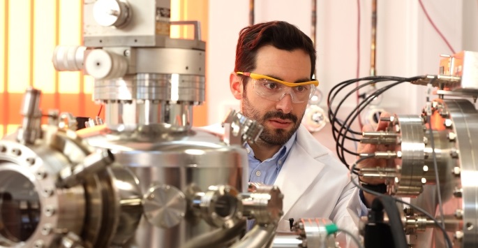 A man wearing goggles sits behind silver machinery with wires.