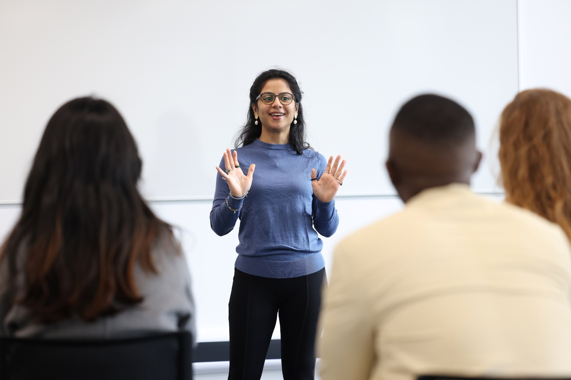 A woman stands at the front of a group of people. Her hands are up and mouth is part way open as if in the middle of delivering a lecture.