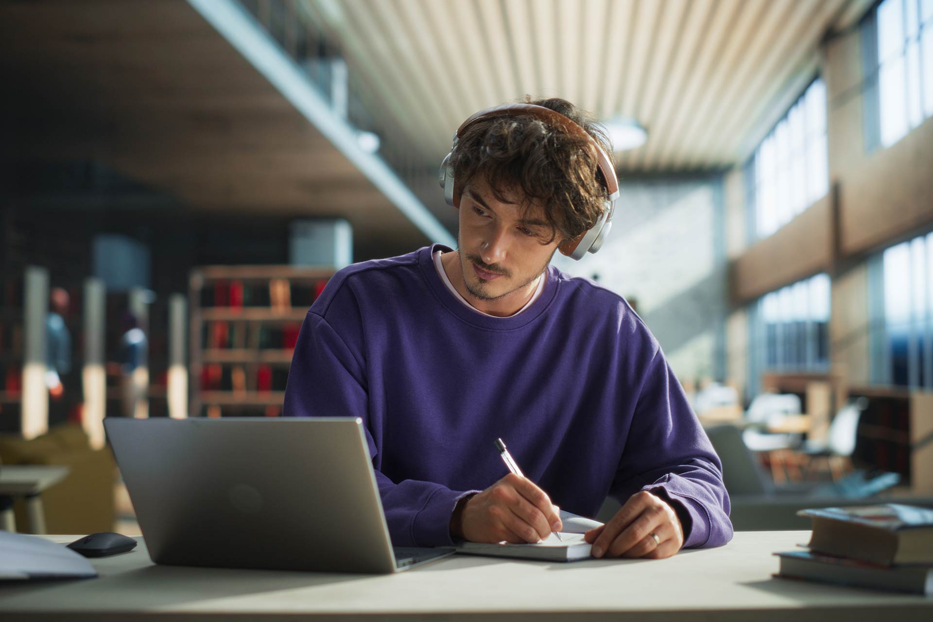 Student wearing headphones writes notes while looking at a laptop screen in a public library.