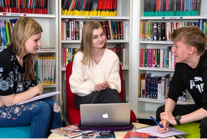 Three students sit together round a table in the library. There are bookshelves behind them. They all have laptops and are engaged in conversation.