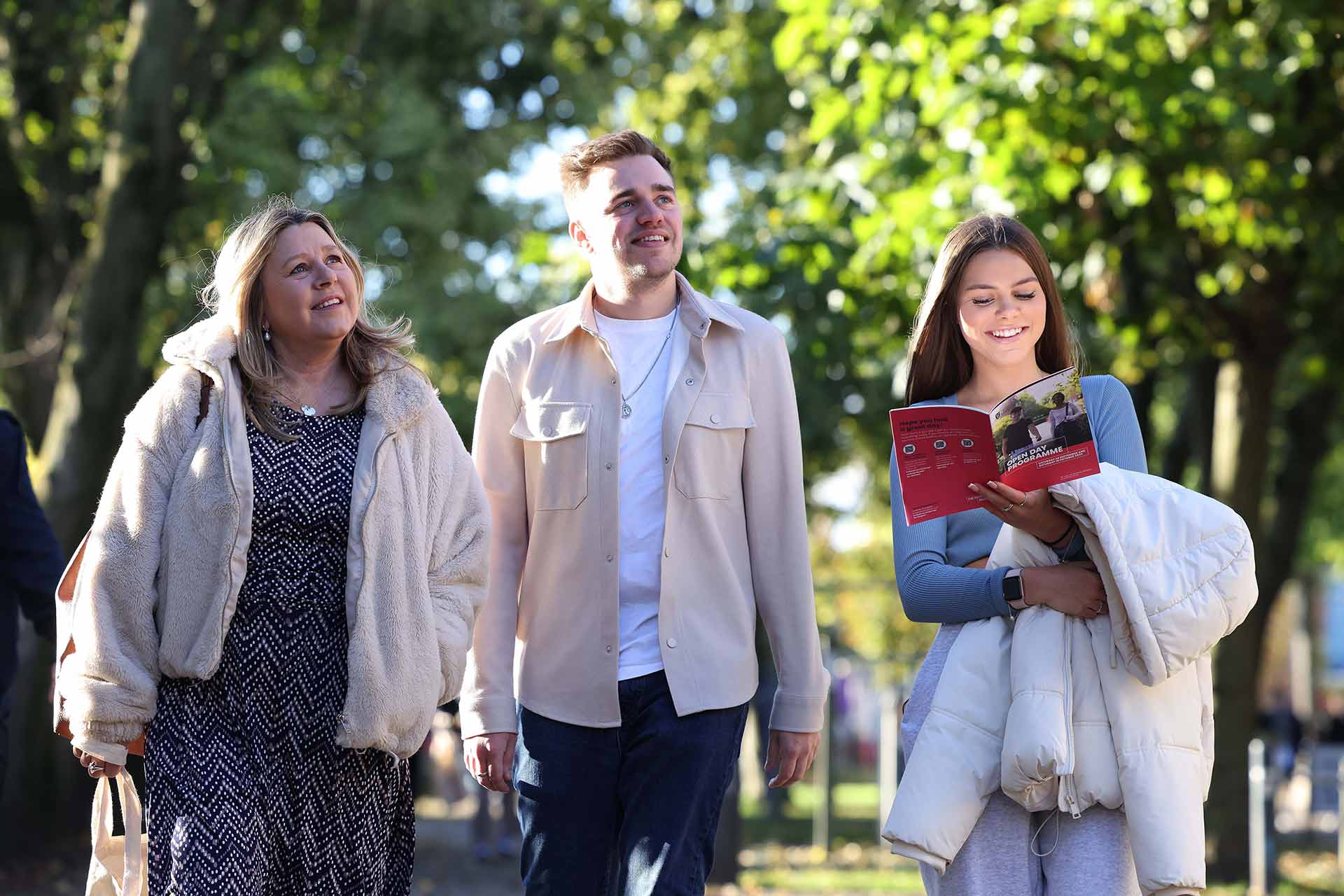 A parent and two prospective students, one male and one female, walk across campus. They are surrounded by trees with green leaves and the sun is shining. The female prospective student is reading an Open Day booklet with a pink cover.