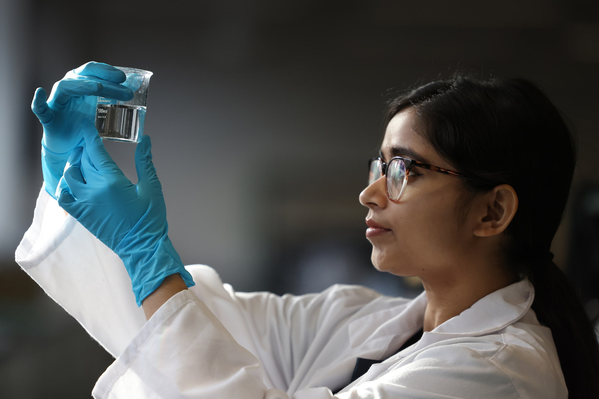 A woman with glasses and black hair holds up and examines a beaker full of clear liquid.