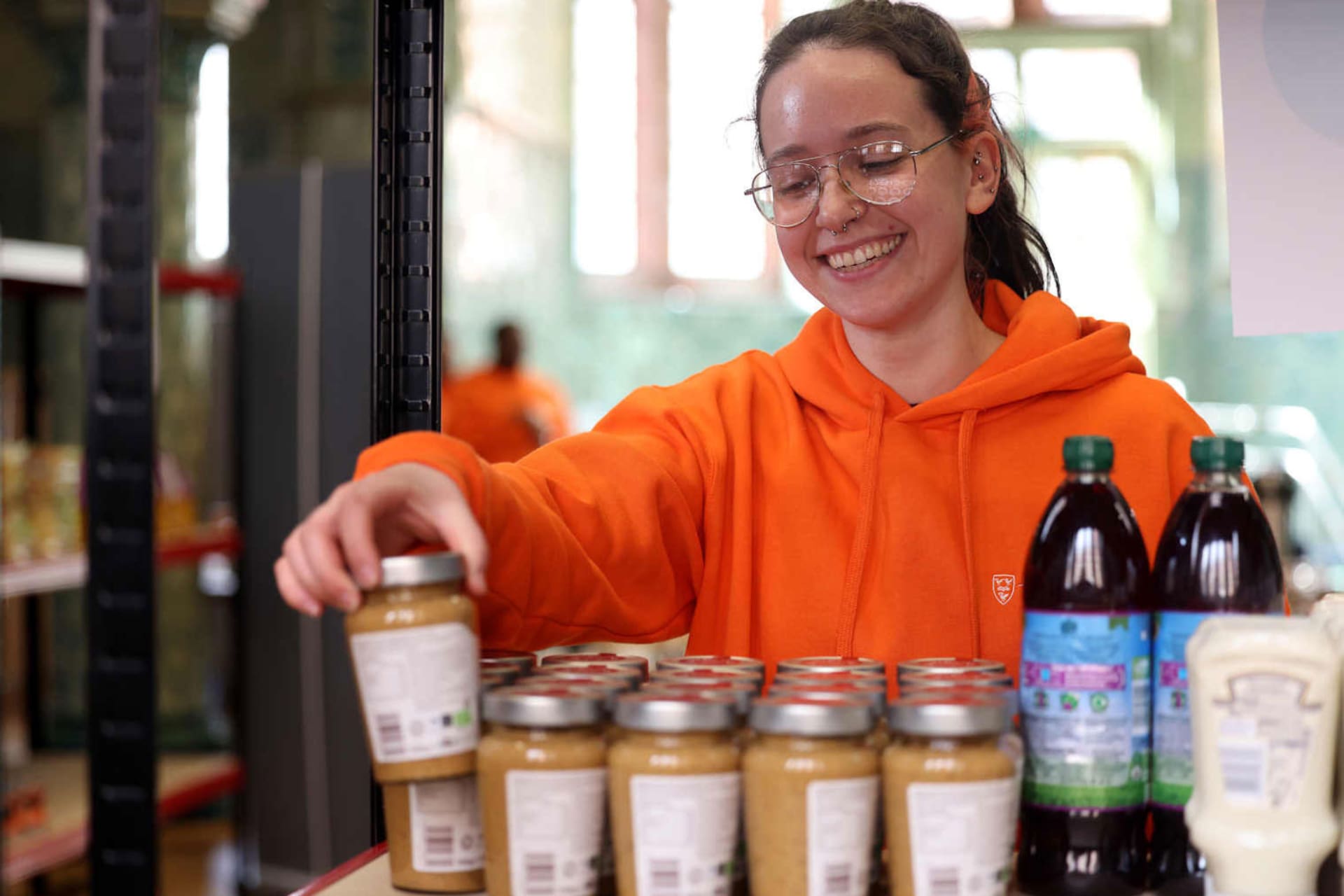 A young woman wearing glasses, smiling as she picks a jar of peanut butter out from a row of jars.