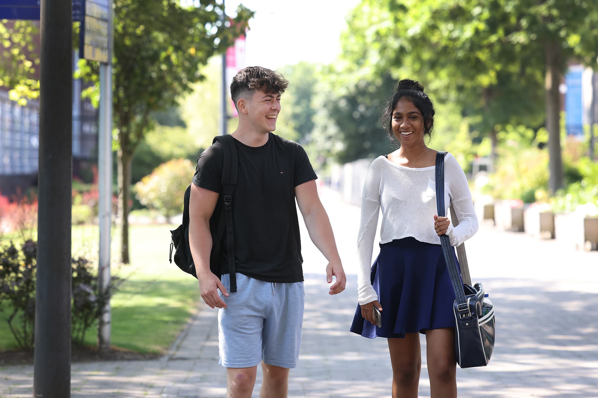 A male and a female student walk together on campus. They are smiling. There are trees in the background.