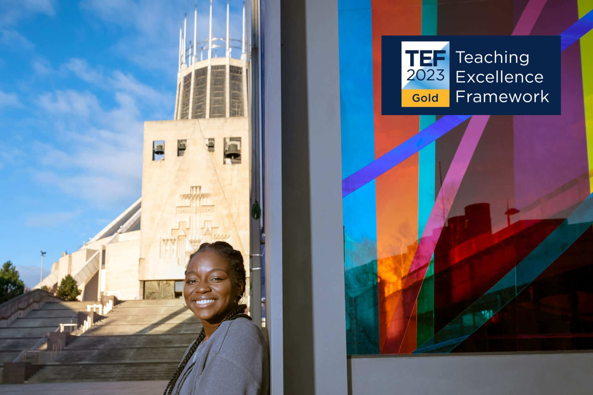 A black female student stands outside the Catholic Cathedral. She is smiling. It is a sunny day with a blue sky. The TEF Gold 2023 logo is in the top left-hand corner.