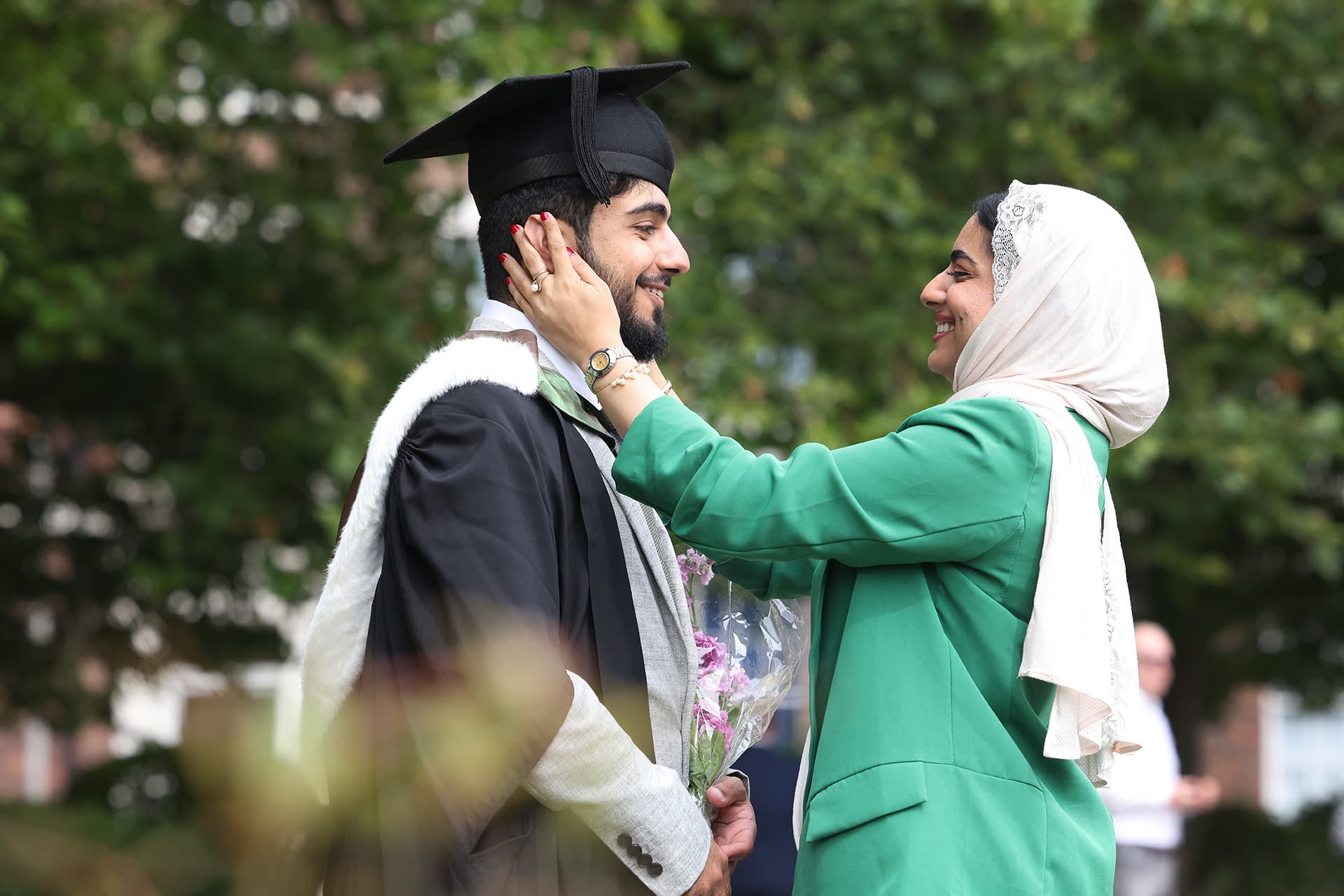 A male wears his graduation cap and gown. He stands facing his mother. She is looking at him and holding his face. She is smiling and wearing a Hijab.