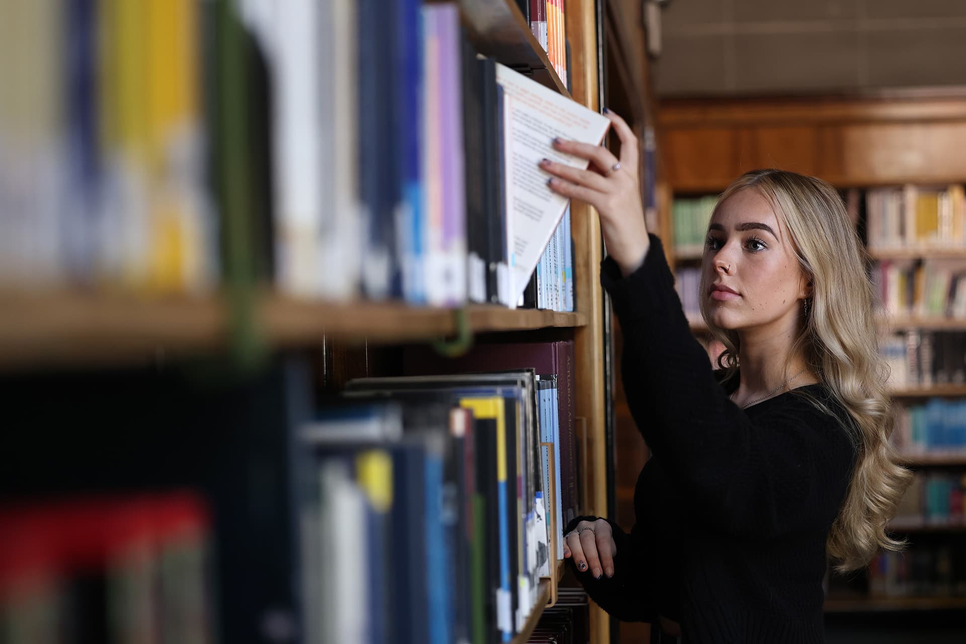 A blonde female student takes a book from a shelf in a library.