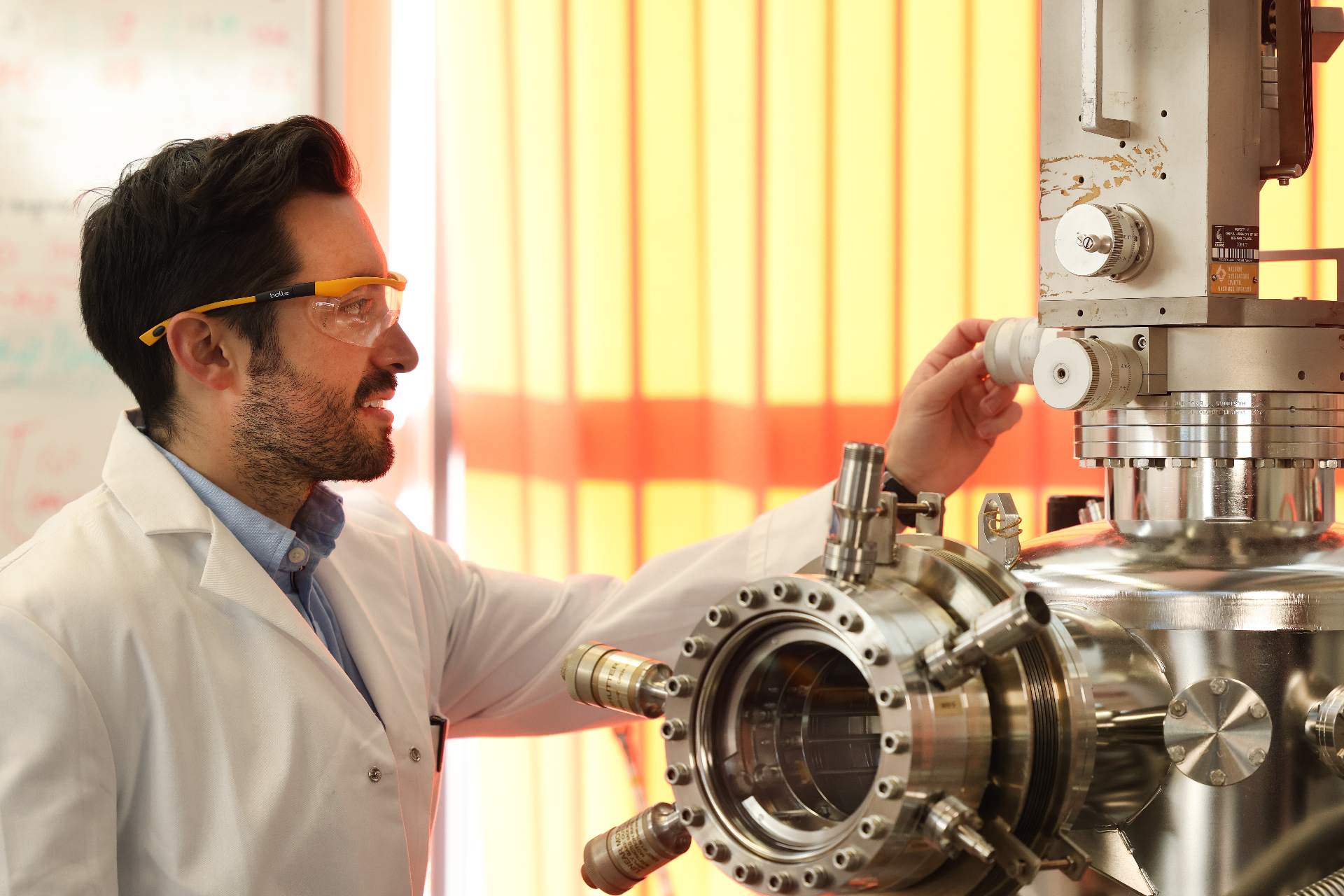A male student works in a lab with equipment. He is wearing safety goggles and a white lab coat.