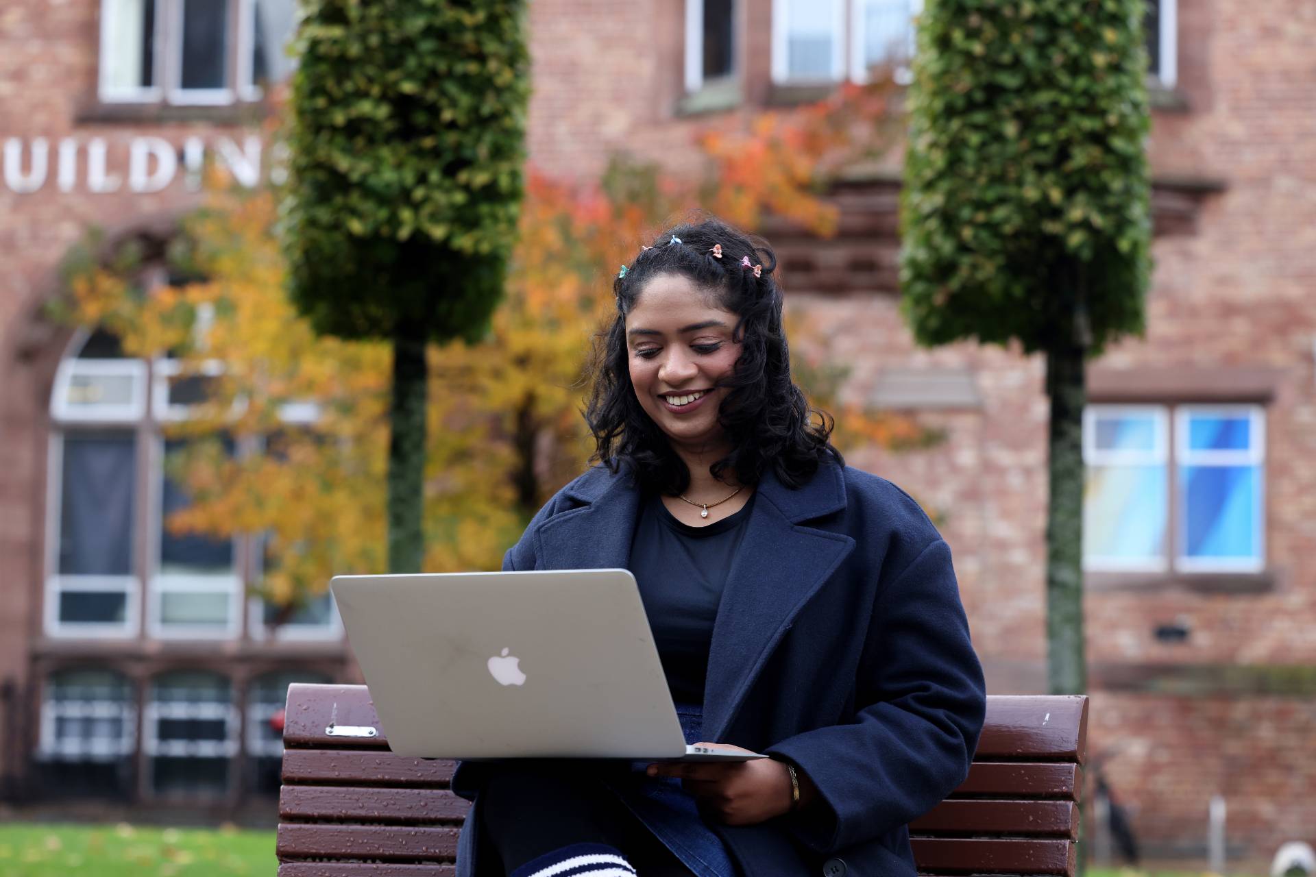 A student sits outside on a bench on campus using a laptop.