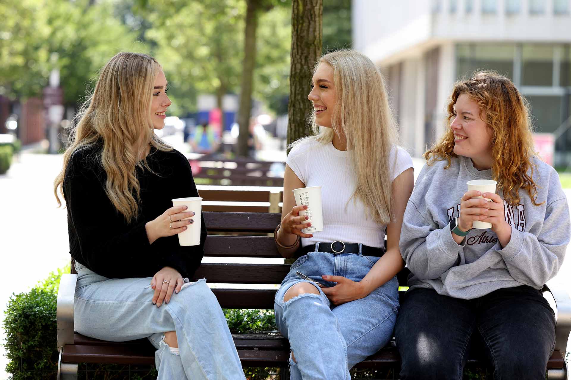 Three students sat on a bench on campus with hot drinks