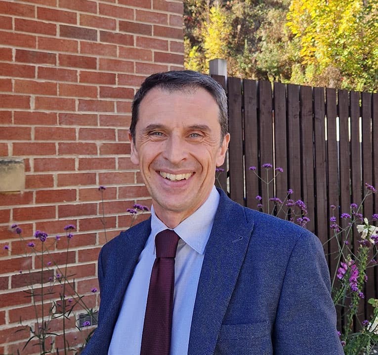 Photograph of Simon Jones. A white male. He is wearing a suit and tie and smiling.