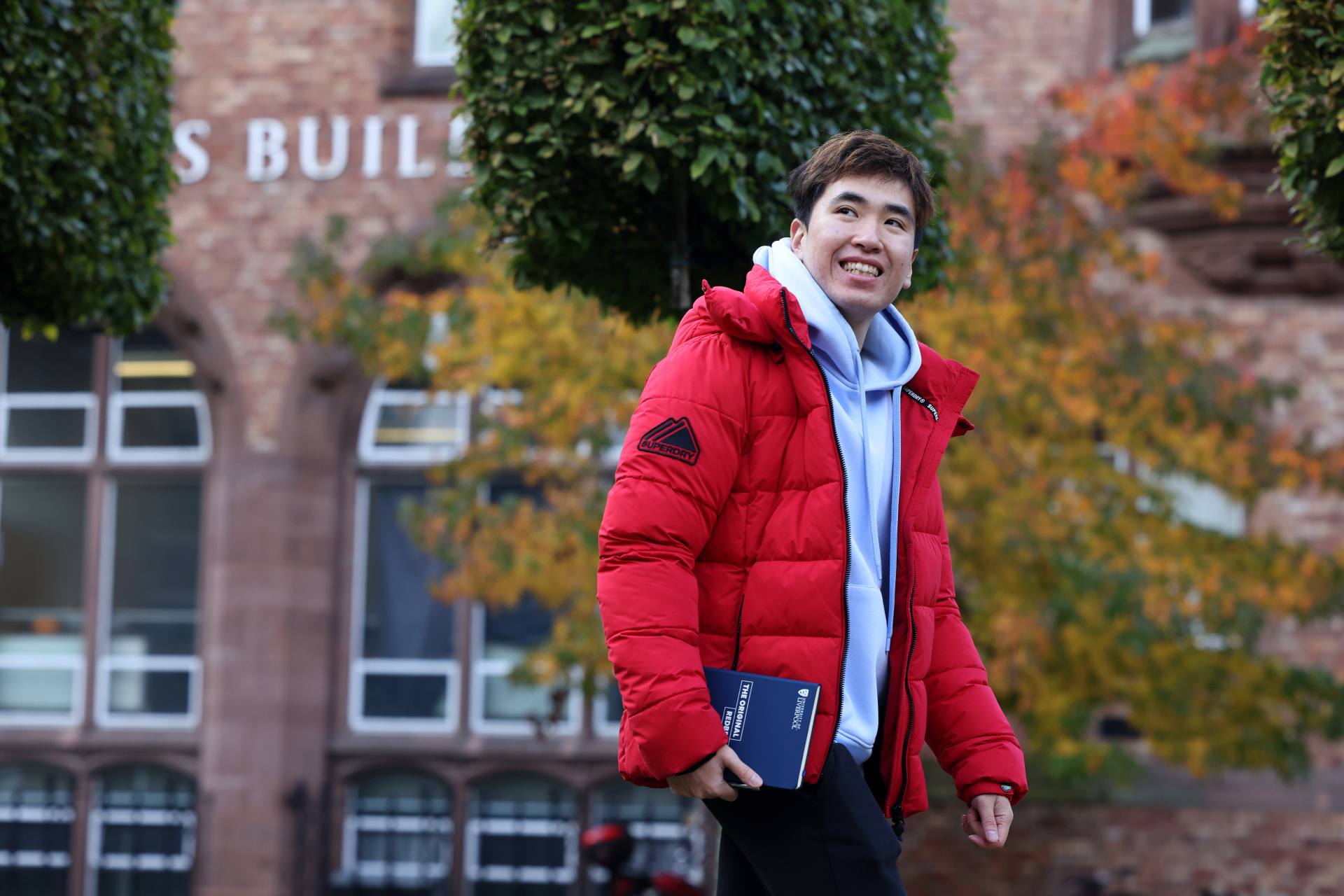 A student walks across campus, holding a book and smiling.