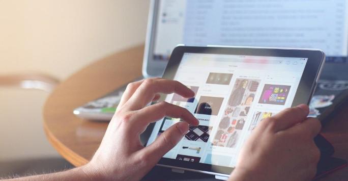A person holding a computer tablet in their right hand whilst using their left hand to navigate the screen. There is also a laptop computer in the background on a table.