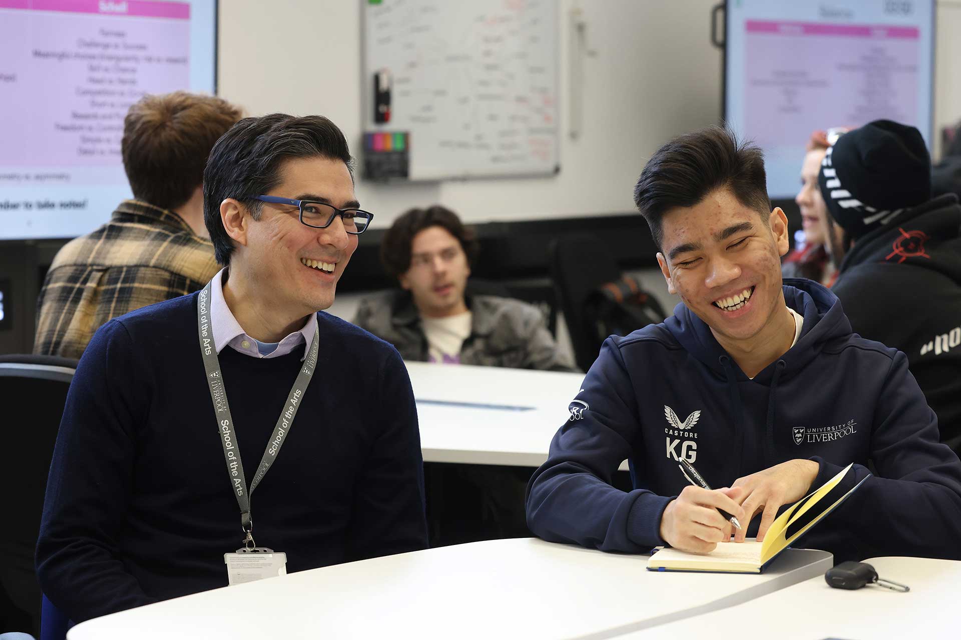 A student makes notes while talking to their tutor in a classroom during a seminar.