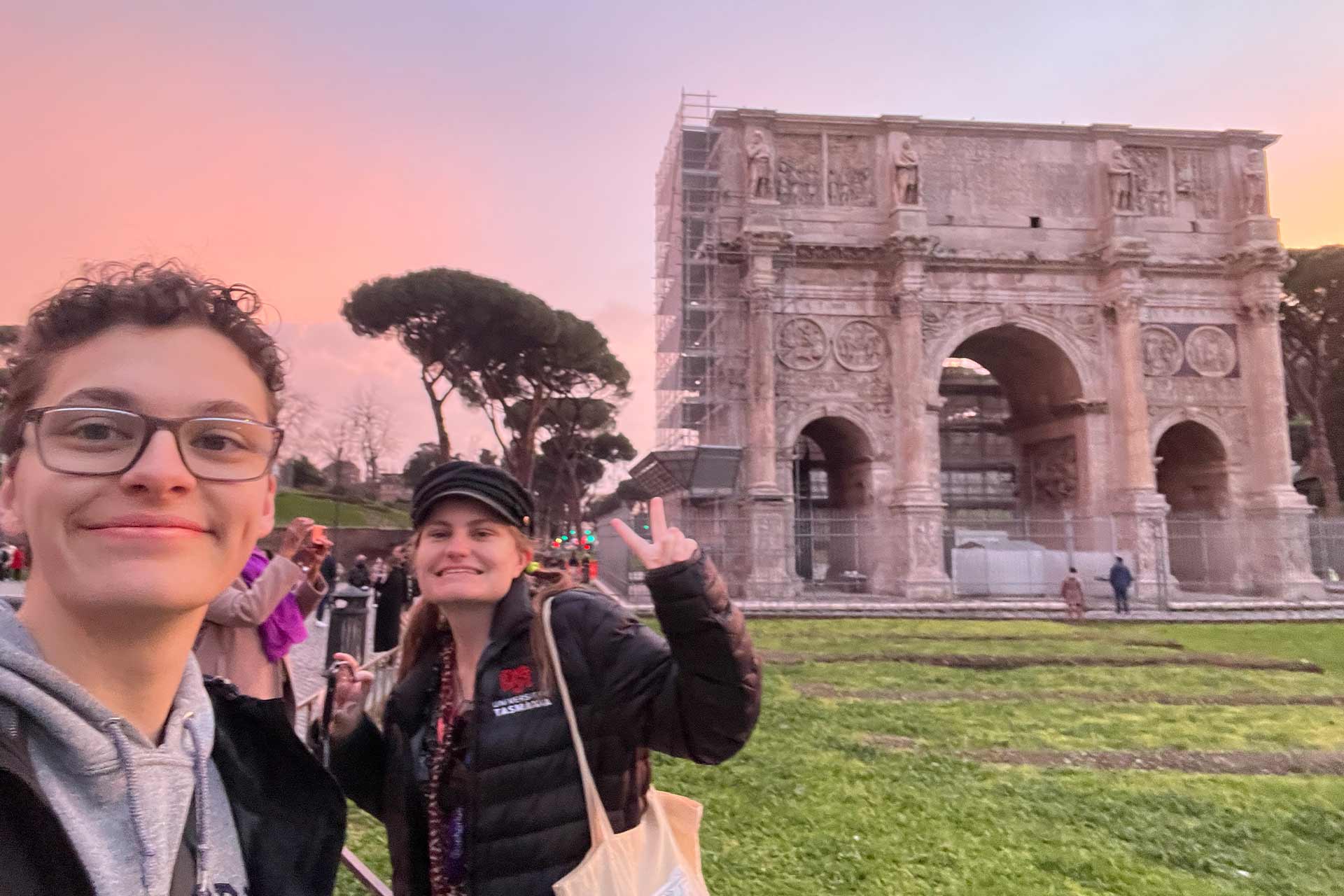 Two young people standing together smiling next to a old stone monument that has three archways in it.