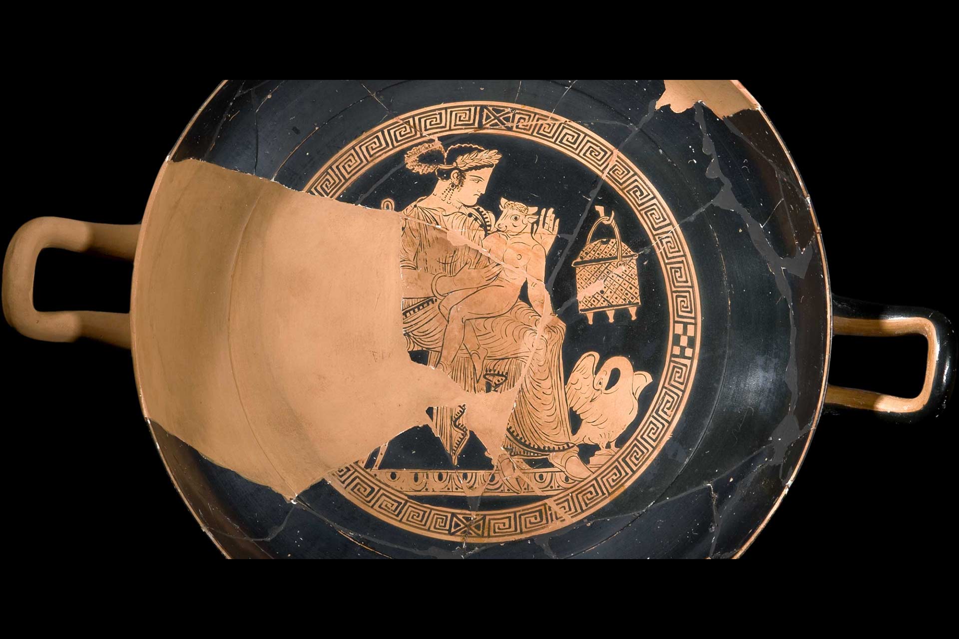 An old worn black and copper plate or bowl with handles with a painted person in the middle holding a small minotaur on their lap