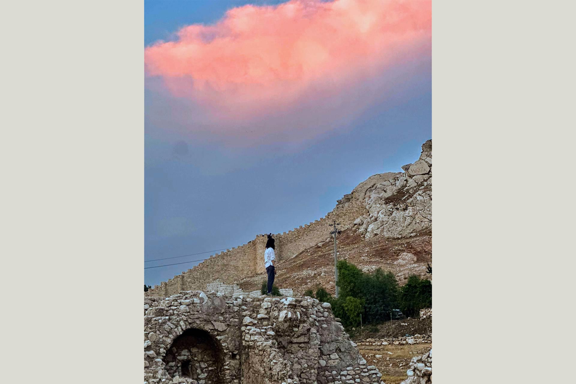 A woman standing facing old gates and stone walls in a rocky hilled area