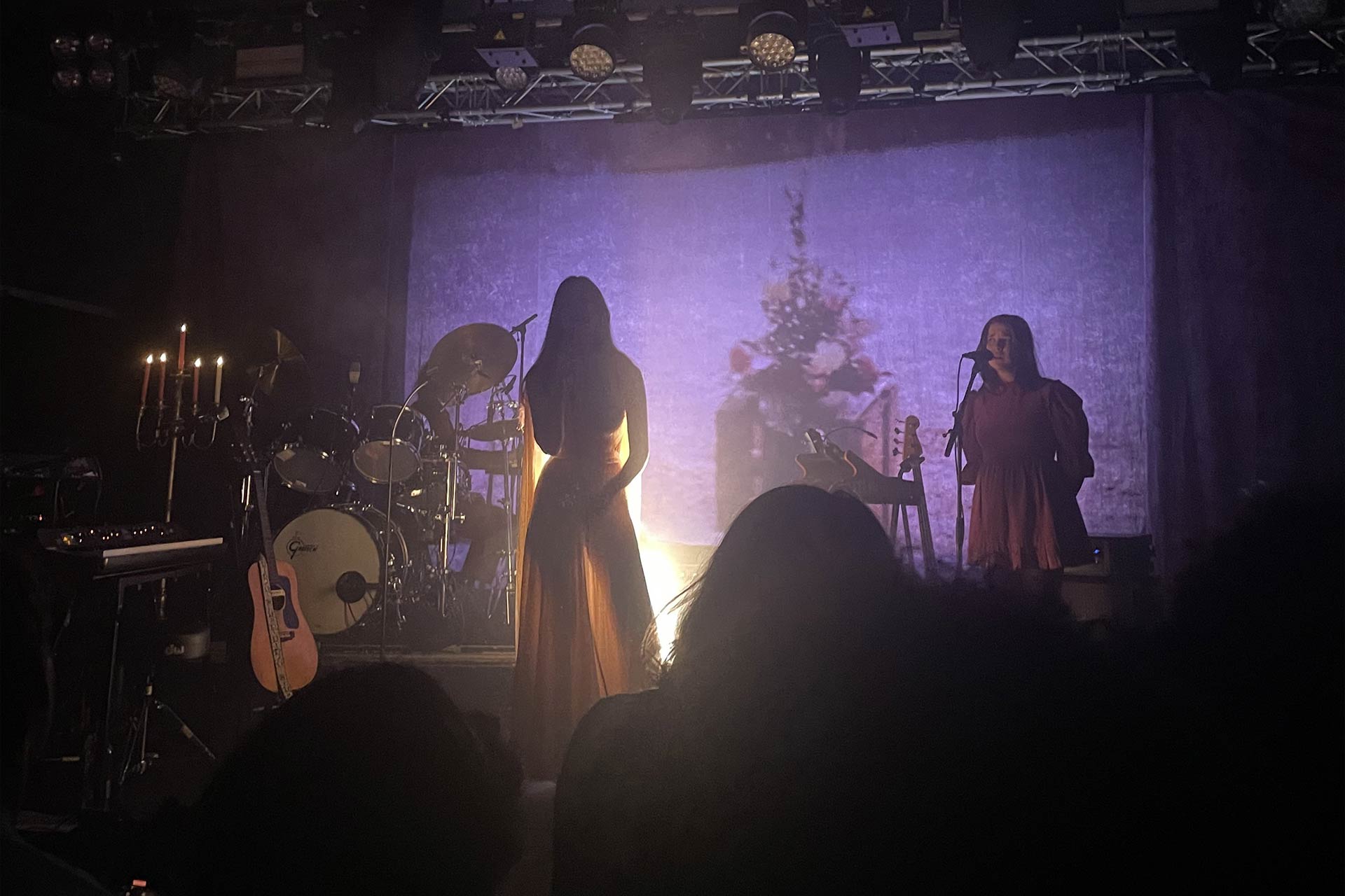 A band on stage, at the centre is a woman in a floaty sheer skirt and the band are dimly lit from behind creating silhouettes