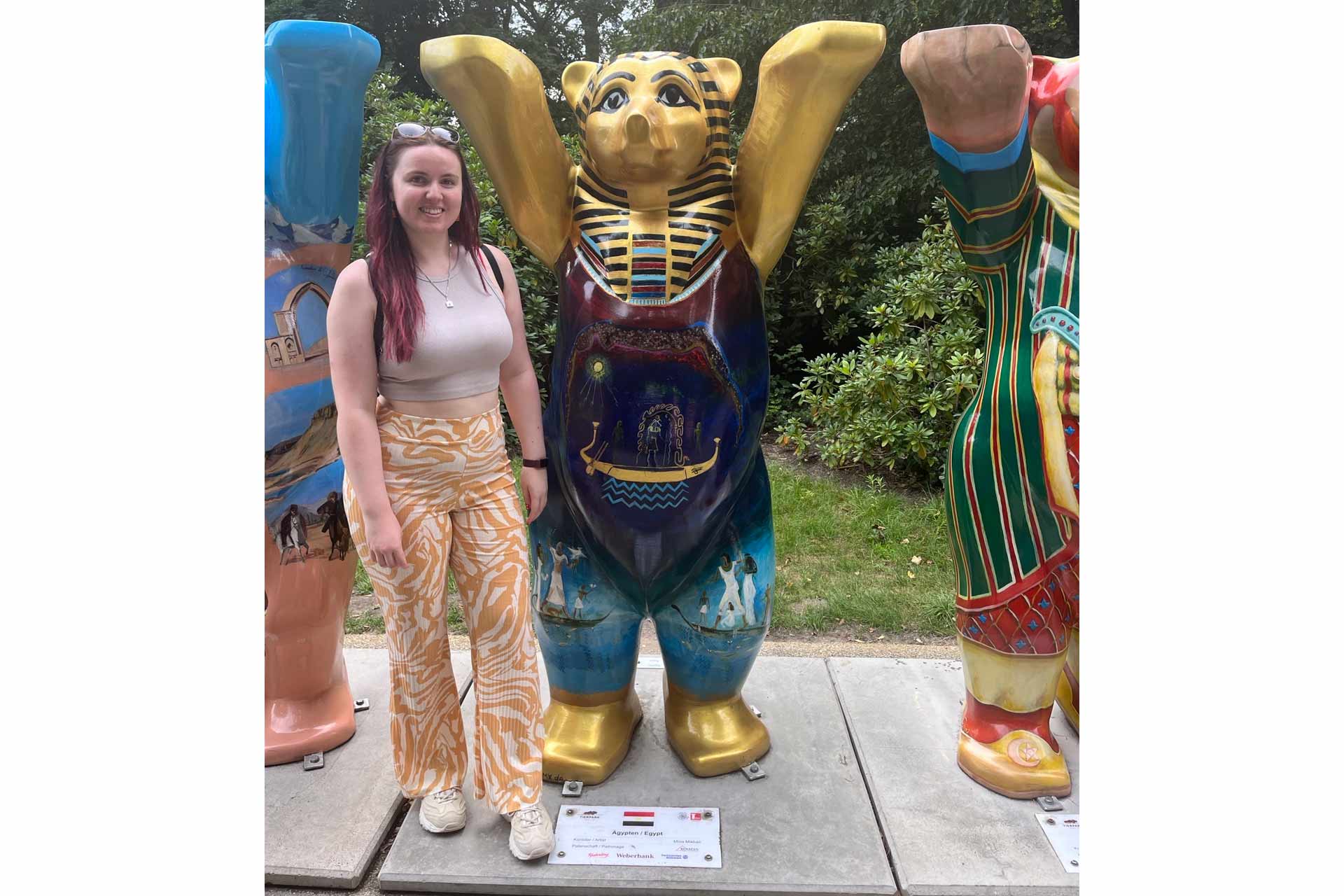A woman with brown hair standing next to a gold statue of a bear that has Egyptian markings on it