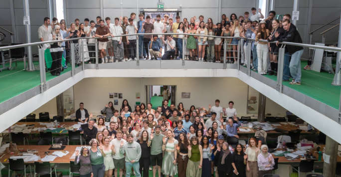 Large group of students on two leverls waving at the camera. The photo is taken inside a large modern hi-tech studio.