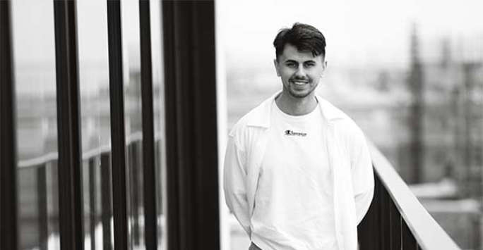 Photograph of a man standing on the balcony of a high rise building, and urban landscape visible below.
