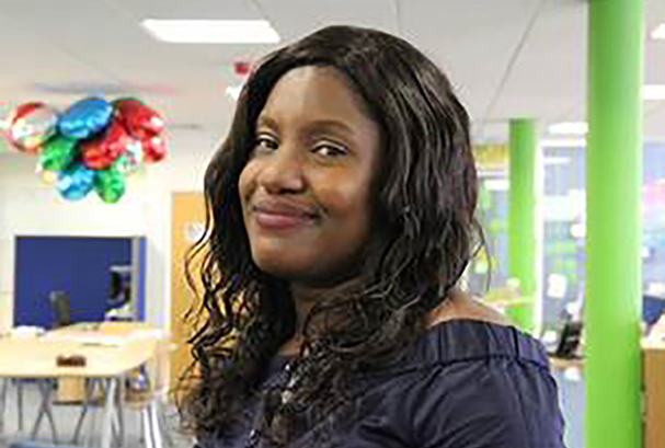 Black woman with long dark hair, wearing a blue top and smiling. She is standing in open plan office with balloons floating on the ceiling