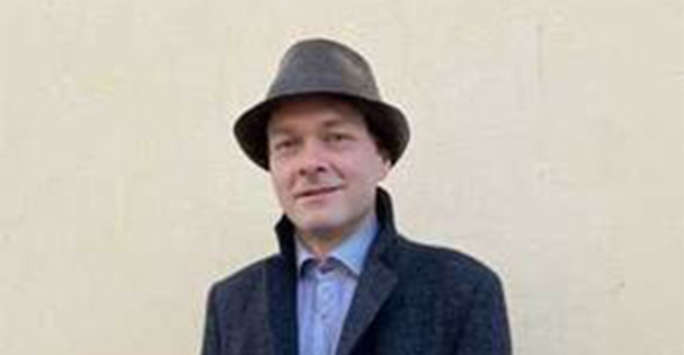 Middle aged white man wearing a Dark blue coat, blue shirt and grey hat standing in front of a featureless wall