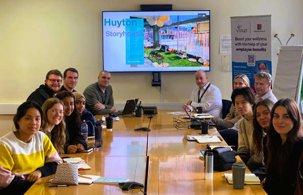 Women and men sitting either side of a long wooden table. At the head of the table is display screen showing a an image of an urban village with the text
