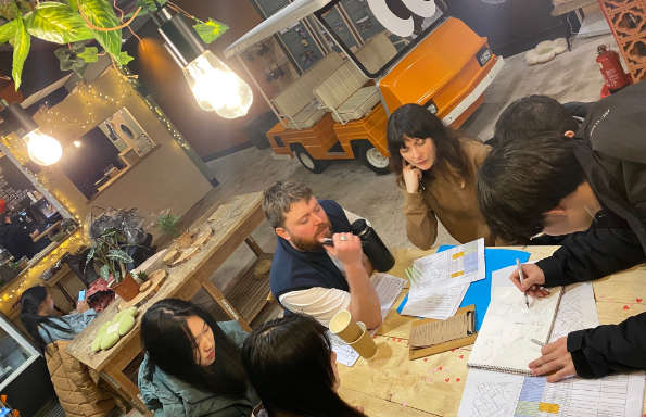 In a spacious cafe men and women sitting around wooden tables, sketching and discussing projects. A golf buggy is in the background.