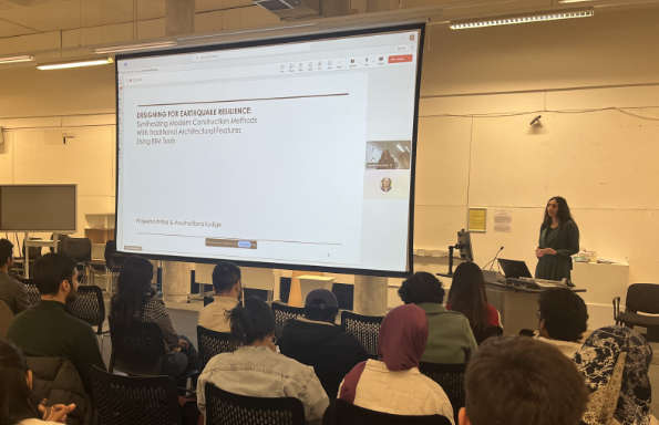 A woman stands by a projection screen speaking. On the screen is the text 