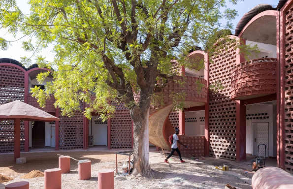 Man walking through a courtyard surround by modernist clay brick built buildings. In the middle of the courtyard is a large tree.
