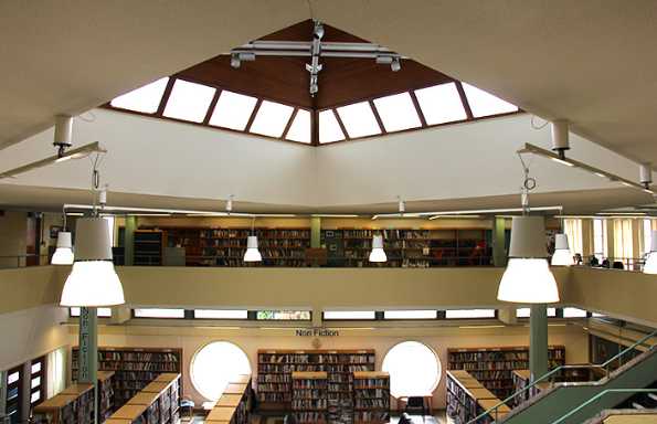 Inside of a 16's library with a pitched floating roof and stairs leading up from the ground floor. Bookshelves line the walls around an atrium.