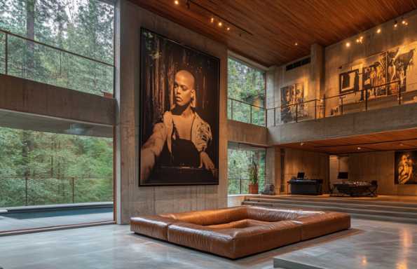 Interior of a modernist building, raw concrete walls and wooden details. In the middle of the room a square leather sofa face a large photograph of an African American woman on the wall.
