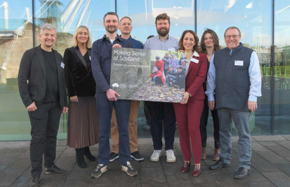 Group of men and women, dressed in a smart casual style, standing outside in front of a glass fronted building. They are holding up a large poster featuring children looking at a stone building with the text 