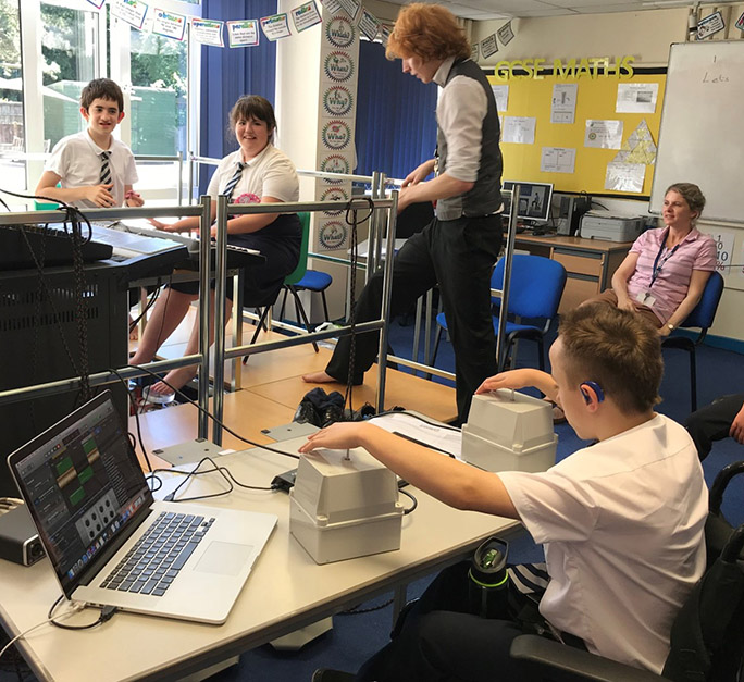 A school classroom, at the back are two pupils sitting in front of keyboards, in the foreground a deaf child with a hearing aid has their hands on two shakers so they can experience sound in a tactile way and participate equally with the other children.