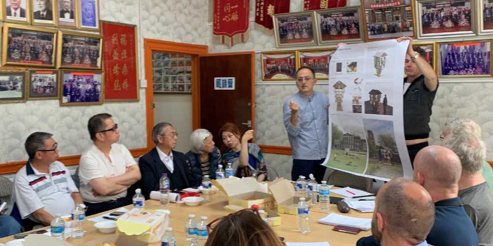 A group of European and Chinese people sitting round a table. At the end of the table two men are holding a design masterplan.