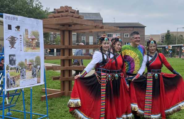Four people, three in traditional Chinese dress stand outside in front of a tall wooden block sculpture.