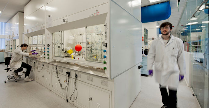 A modern laboratory with a researcher at a workstation and another walking by, both in lab coats and gloves.