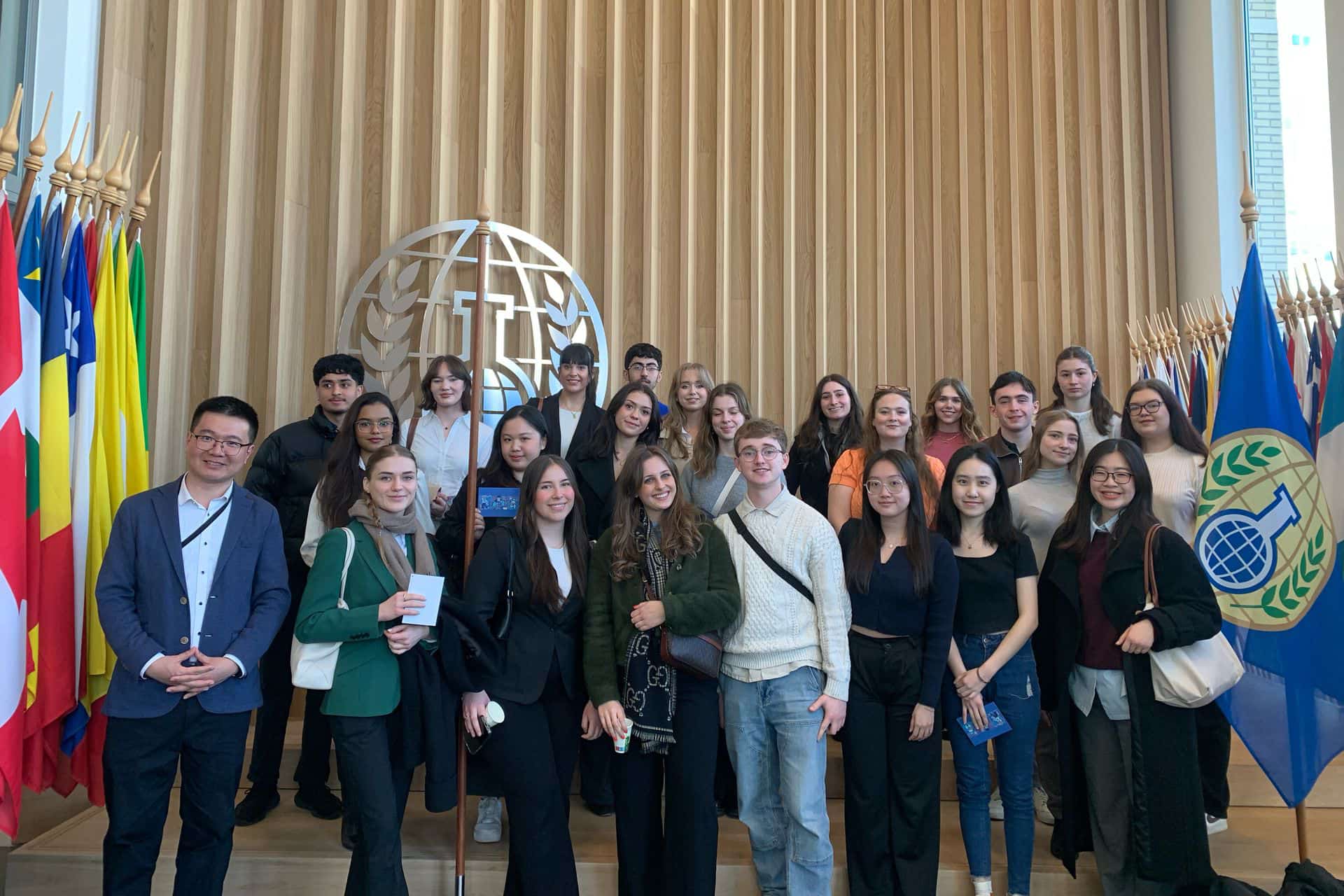 The large group of students from the study trip stand together surrounded by international flags.