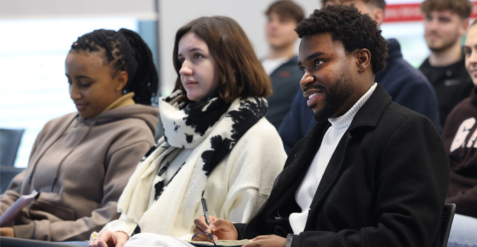 A row of students sit facing towards the front of the room.