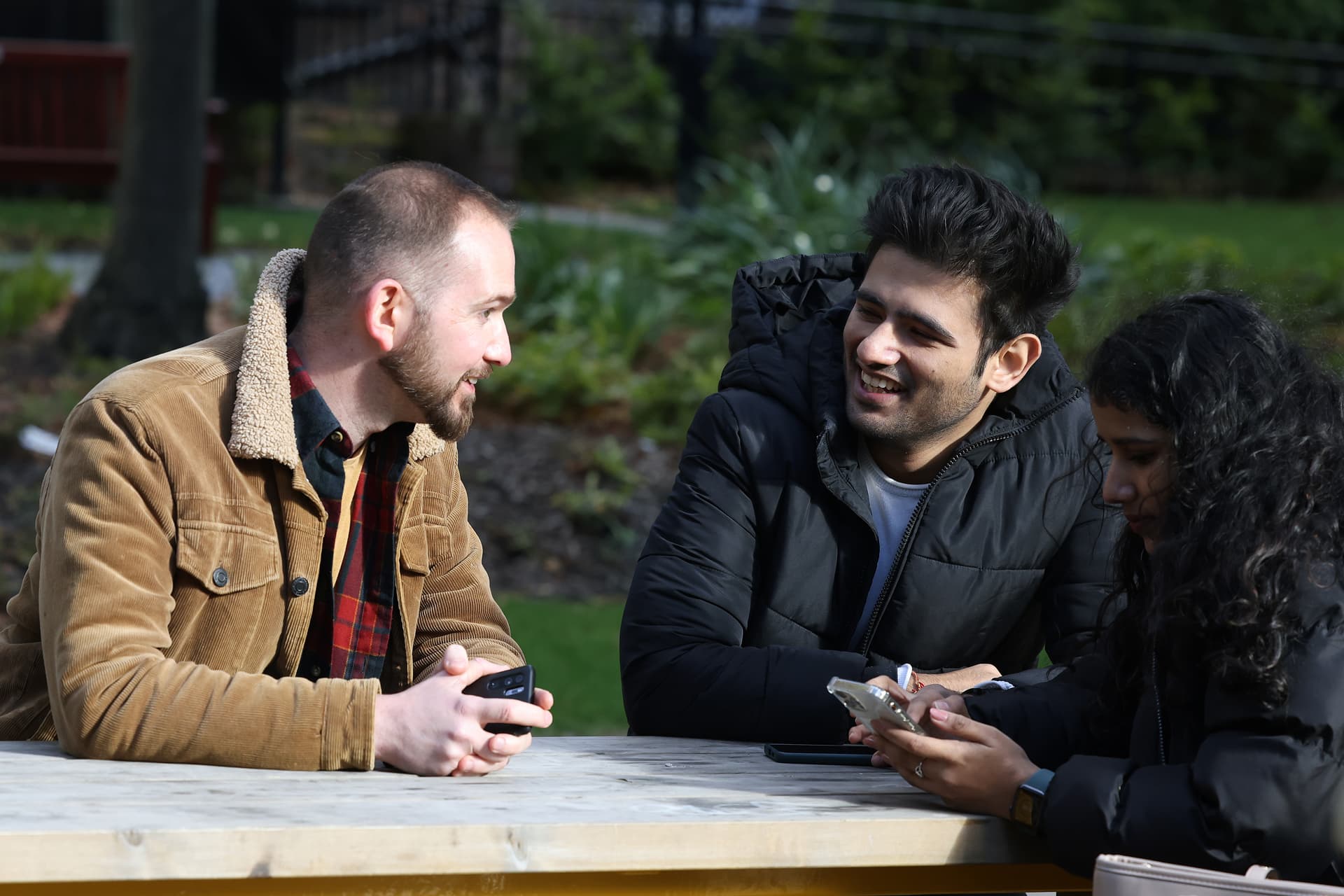 Three students sit around a bench together. Two students are in conversation whilst the other is looking at her phone.