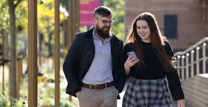 Two students walk together through campus whilst looking at the phone in one of the students hands.