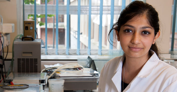 A student wearing a lab coat stands in a lab.