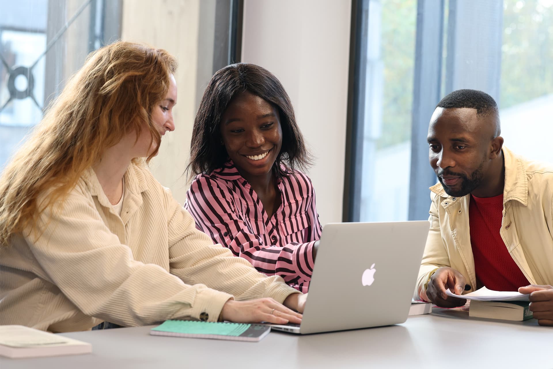 Three students sit together at a table in a study space, focused on the screen of a shared laptop.