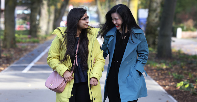 Two students walking through campus, walking along a path that is lined with trees.