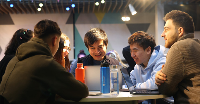 A group of student sit around a table together. They are looking at a a laptop.