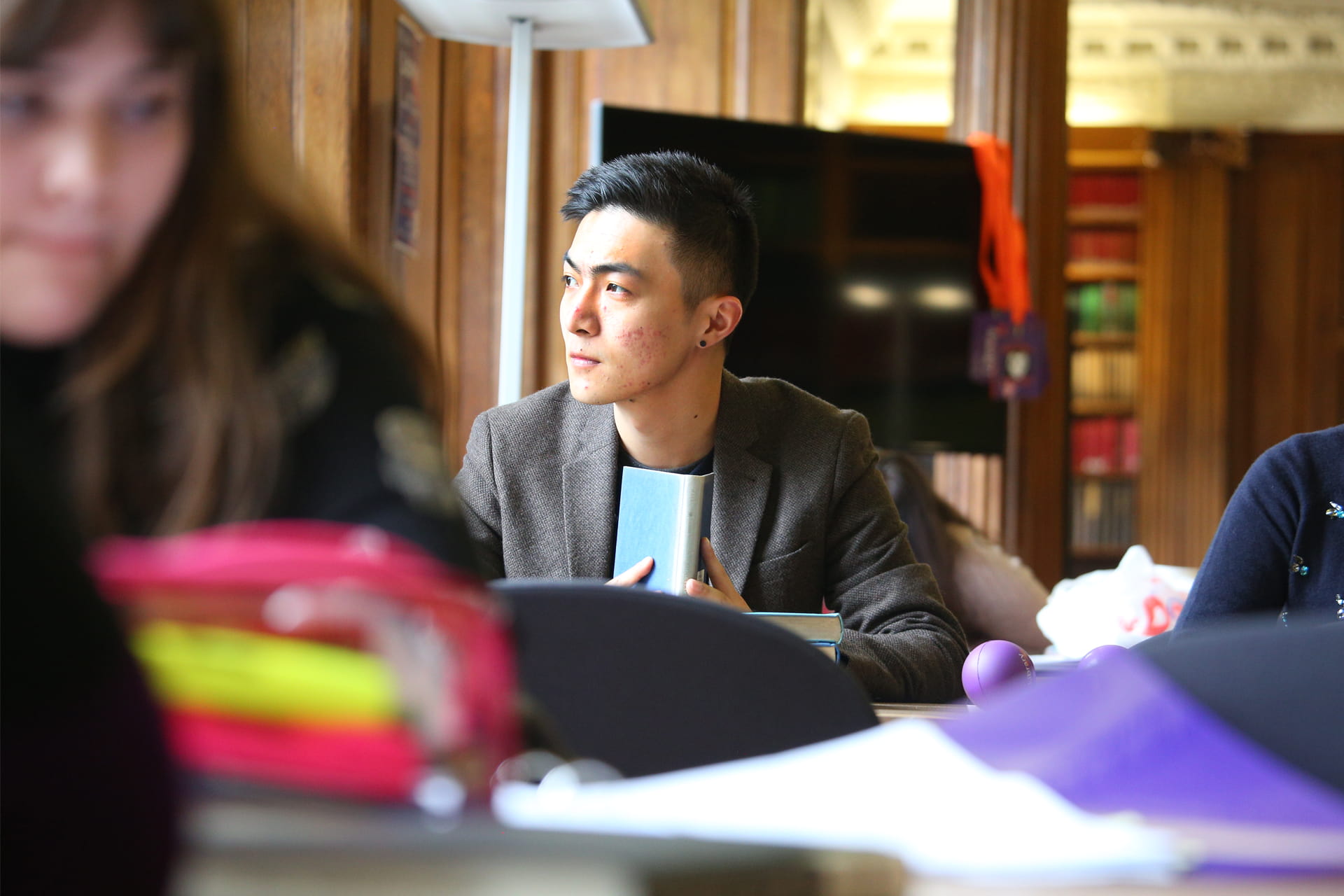 A students sits at a desk holding a book whilst gazing out of the window.