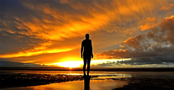 An Antony Gormley statue of a male figure stands on a beach, gazing out at the sea. Behind it, a bright yellow sun sets behind the clouds.