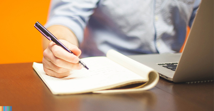 A person writing in a notebook with a laptop in front of them